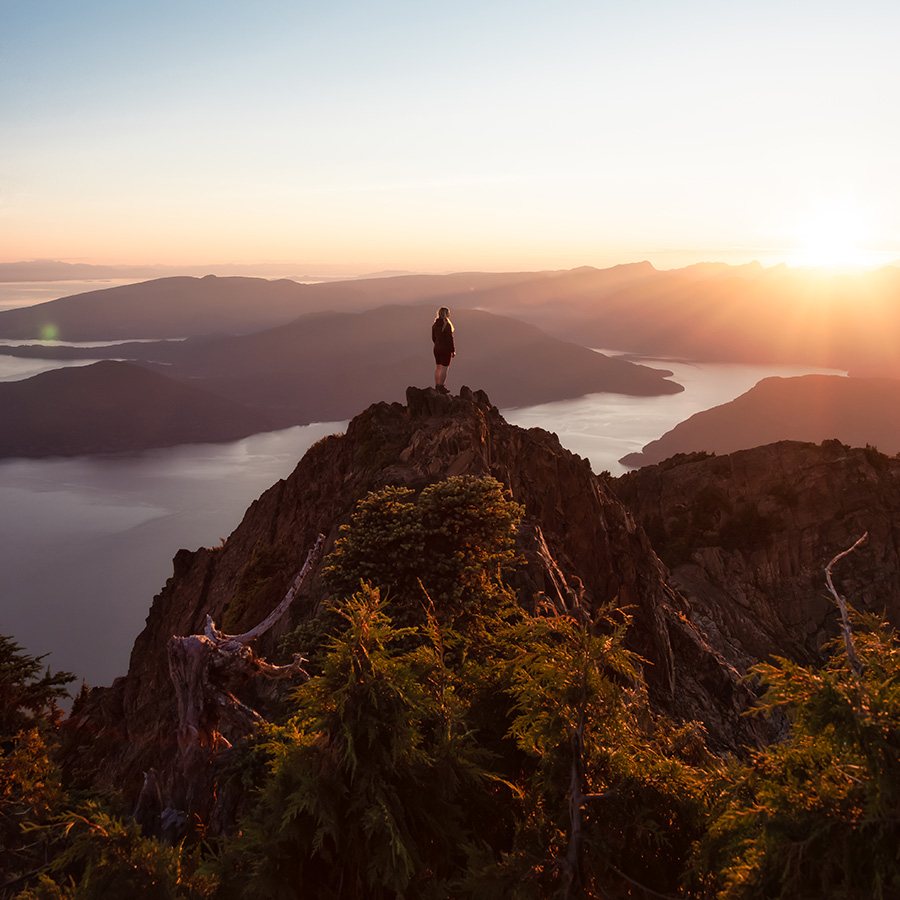 a women standing on a mountain peak, overlooking a tranquil lake surrounded by mountains during sunset, conveying a sense of peace and adventure